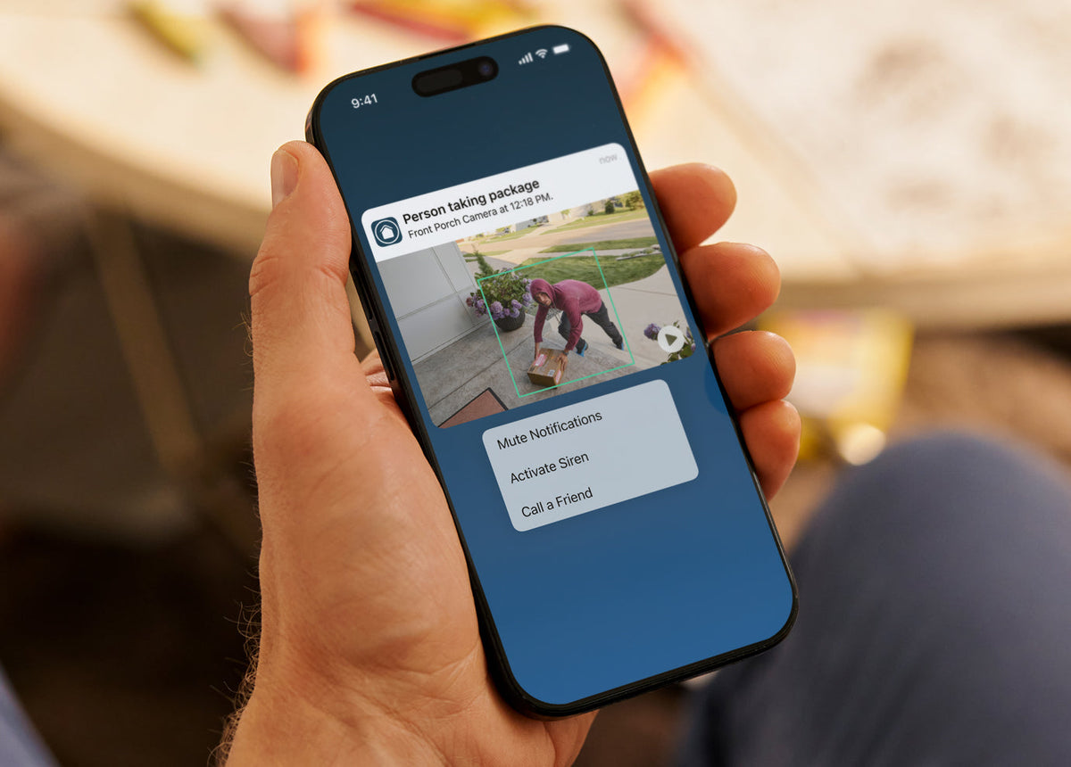 Hand holding a smartphone displaying Arlo Secure app with a person taking a package, blurred background