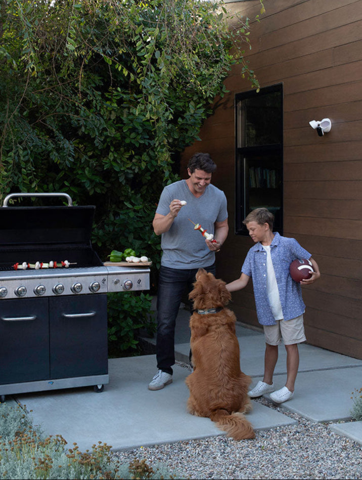 A father, son, and dog outside BBQing with the Arlo Wired Floodlight mounted on the outside of a house
