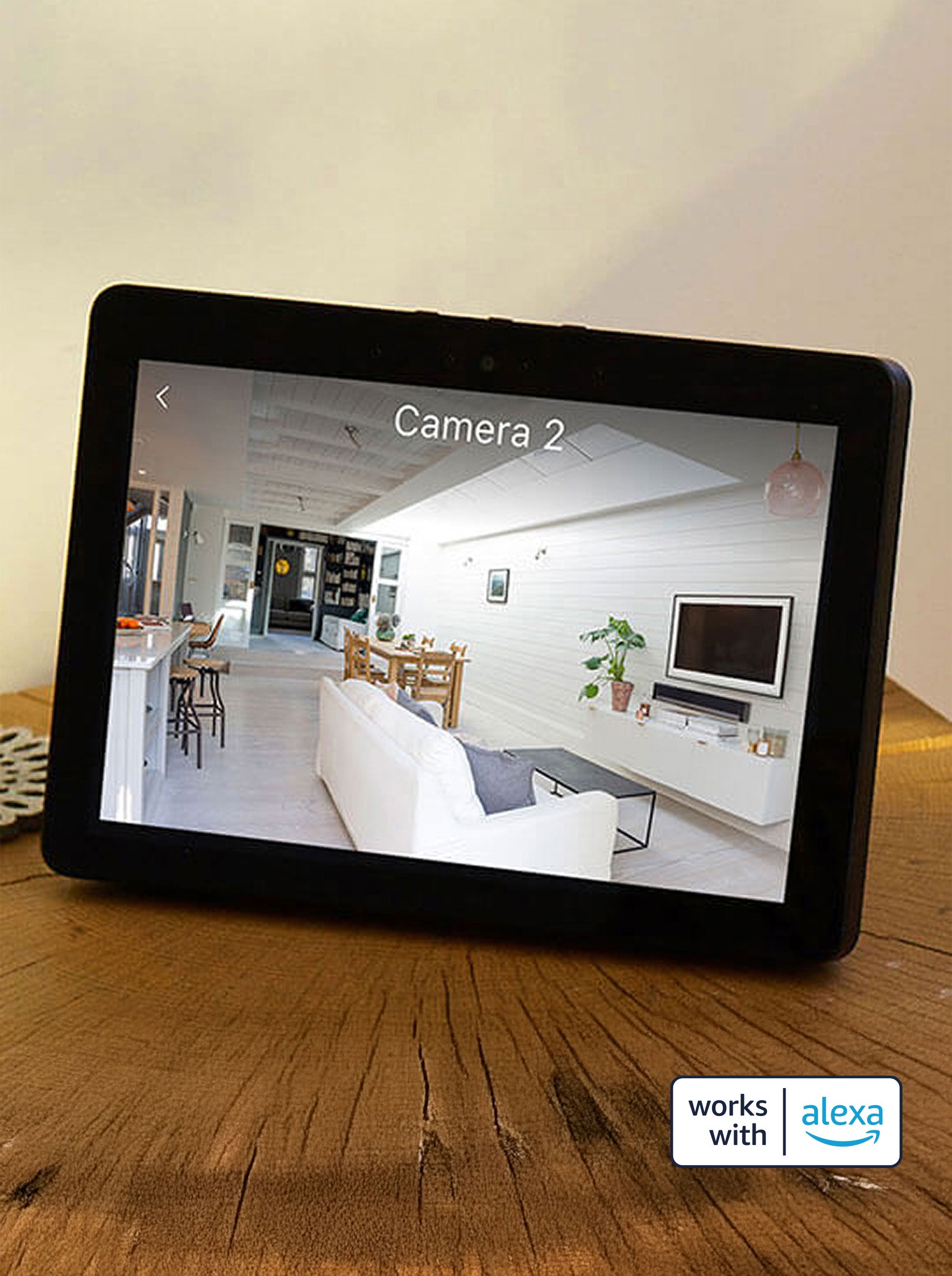 A tablet screen displays a live feed from an Arlo security camera, showing a modern, well-lit living room and kitchen area. The camera is compatible and pairs with Amazon Alexa for convenient visual & voice control.
